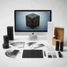 A desktop computer displays a 3D geometric cube for Packaging Design. Around the monitor are packaging mockups, color swatches, a graphics tablet with stylus, and samples—essential tools for graphic or product design on a clean white surface.