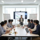 During a Project Management Training session, a woman presents a chart on a large screen to eight colleagues seated around a table with laptops, papers, sticky notes, and coffee cups; diagrams are on whiteboards.
