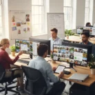Four people collaborate on Stock Photo Curation, discussing images displayed on large monitors in a bright office with windows, plants, whiteboards filled with notes, laptops, and notebooks.