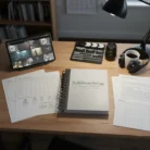 A desk features the “Video Scripting & Storyboarding” spiral-bound notebook, along with shot lists, schedules, and character sketches. A tablet, camera lens, and headphones nearby suggest active video production and story planning.