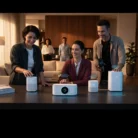 Three people film Corporate Training Videos around a kitchen island with sleek white smart devices, while two others watch from a modern living room with warm lighting and wood and neutral décor.