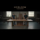 Two men in dark shirts stand on either side of a large wooden table in a bright workshop, evoking the style of Corporate Training Videos. At the top, “HEIRLOOM HANDCRAFTED” is centered.