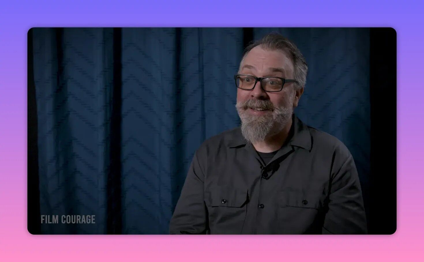 Interview shot of a bearded man with glasses smiling kindly while seated in front of a blue curtain backdrop.