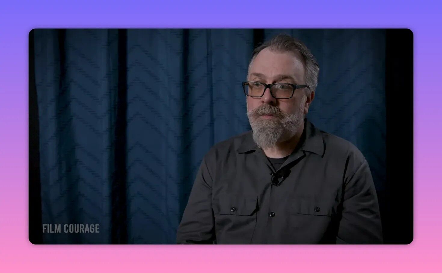Bearded man in an interview looking thoughtful and reflective against a blue curtain background.