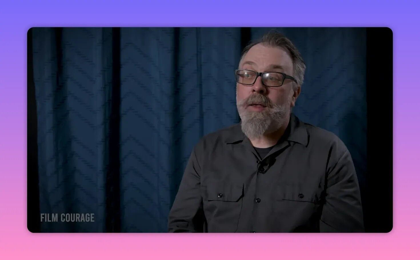 Well-lit interview portrait of a bearded man seated against a blue curtain, speaking thoughtfully.