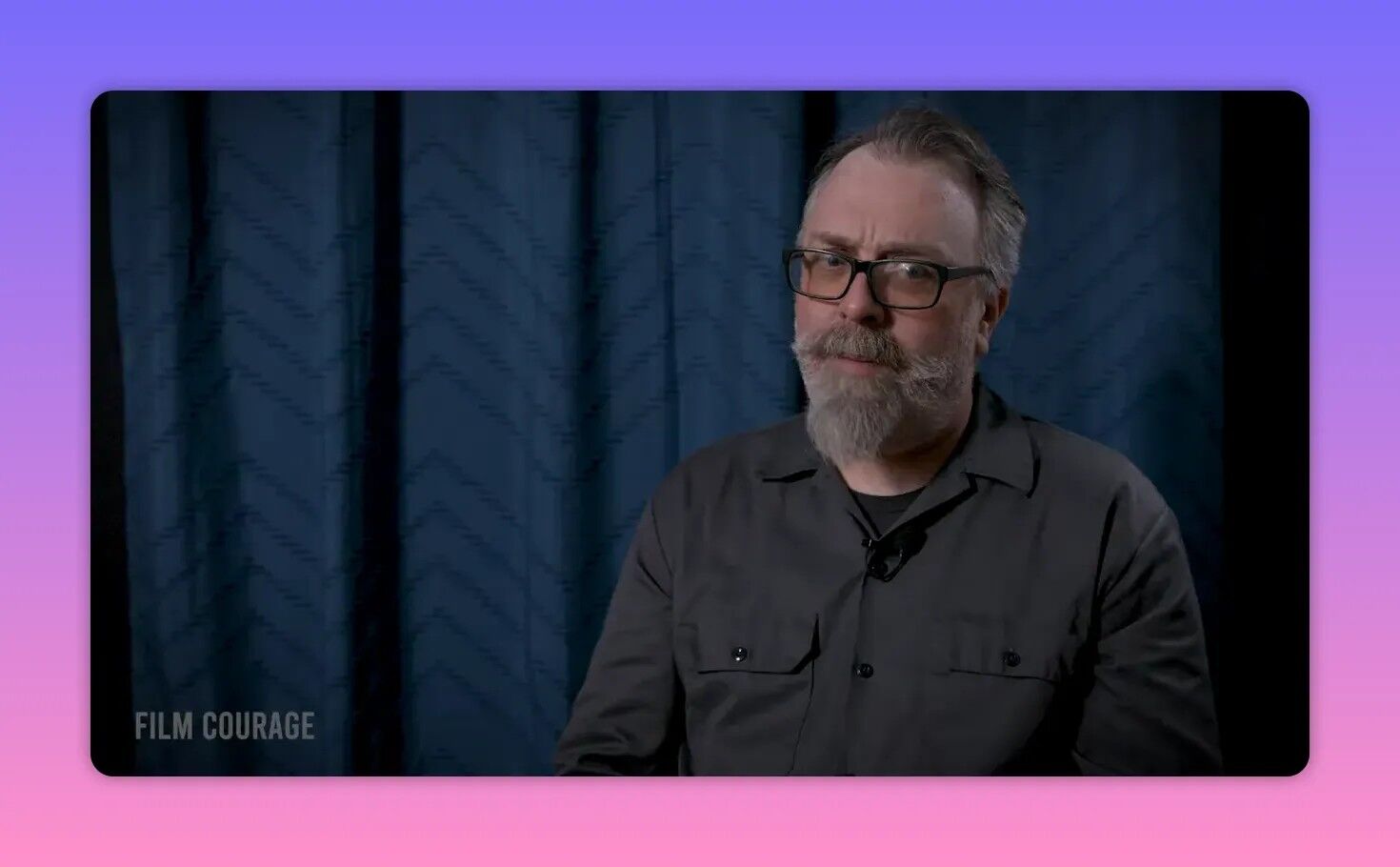Bearded man with glasses in a clear, well-lit interview shot against a blue curtain, thoughtful expression.